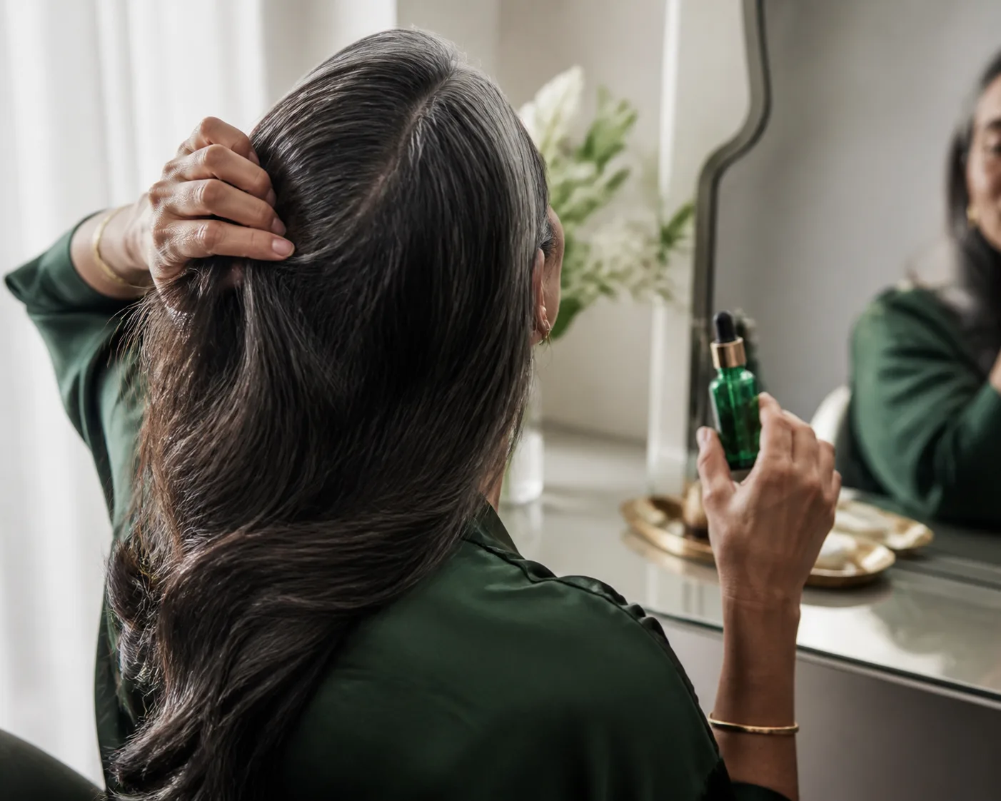A woman parting her silver-streaked hair at a vanity mirror holding an emerald dropper bottle
